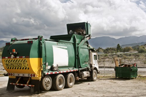 A large green waste collection truck with a black and yellow caution sign at the rear, parked on a dirt surface with a green waste skip nearby filled with mixed debris. The truck has a hydraulic lifting mechanism at the back, partially raised, indicating it is in the process of collecting or emptying waste. The scene is outdoors with a cloudy sky overhead, and distant mountain ranges and sparse vegetation are visible in the background, suggesting a rural or semi-industrial area in the vicinity of St Johns Wood or a similar location. The setting appears to be a waste management site or disposal yard, consistent with rubbish removal services offered by companies like Office Clearance St Johns Wood.