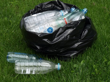 A black plastic rubbish bag sits on a grassy area, filled with and containing several clear plastic bottles with blue caps, some of which are spilling out onto the grass. The bags appear to be loosely tied and are situated outdoors, possibly on a driveway or garden space in St Johns Wood, London. The bottles vary in size, with some fully visible inside the bag and a few resting on the ground nearby. The grass beneath is green and evenly maintained, providing a natural background that contrasts with the black bag and transparent bottles. This image reflects typical rubbish disposal or collection of plastic waste, consistent with rubbish removal services offered by companies like Office Clearance St Johns Wood, within local residential or commercial environments in the area. The lighting suggests daylight, and no additional equipment or tools are visible in the scene.