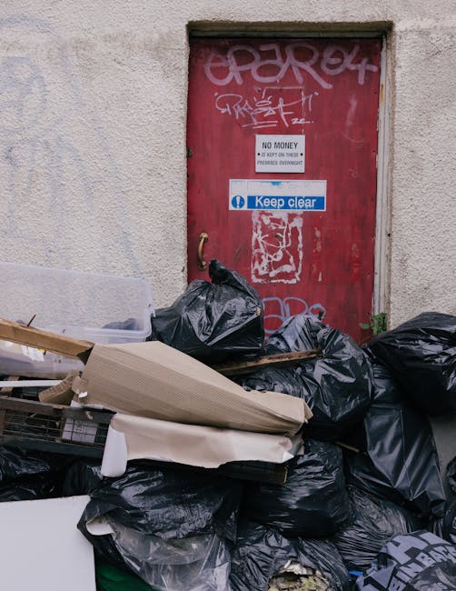 A photograph showing a collection of various-sized waste removal bins and bags positioned on a paved driveway in an outdoor setting. The bins include a large, green, plastic container with a textured