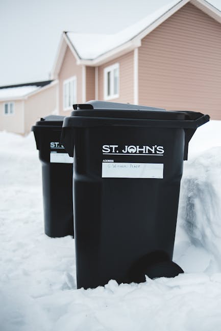 A black wheeled rubbish bin positioned on a grassy outdoor area, filled with garden waste such as dried grasses, leaves, and plant stems. The bin has a closed lid with some garden debris resting on to