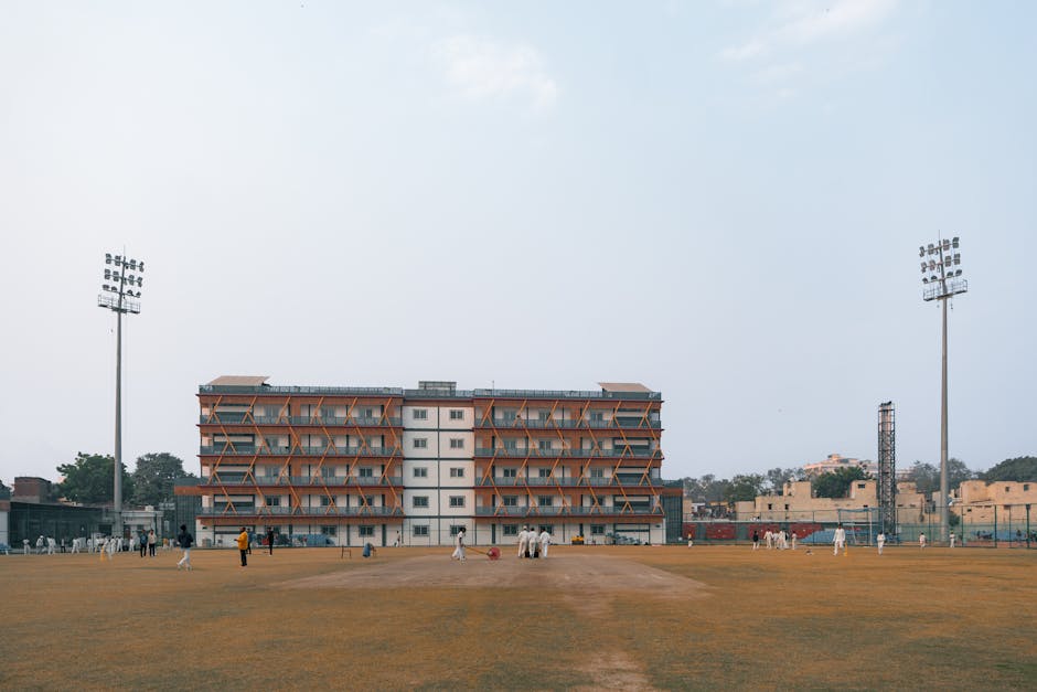 A small brick building with a peaked roof, situated on a grassy sports field, features a large, black rolling shutter door with signage above it displaying the names 'Batsman' and 'Total Batsman'. The