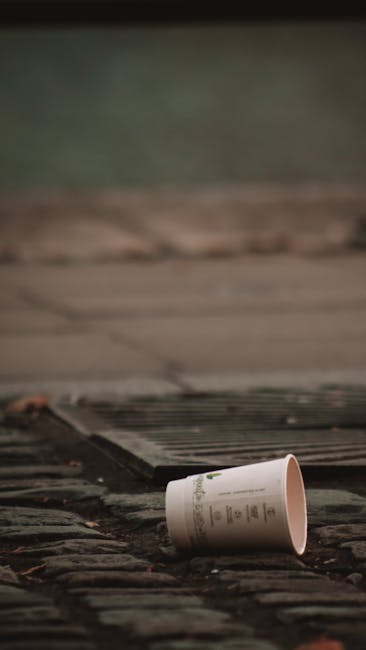 A close-up view of an overturned paper coffee cup lying on a weathered cobblestone pavement, with the cup positioned towards the right side of the image. The cup is white with printed text and logo, p