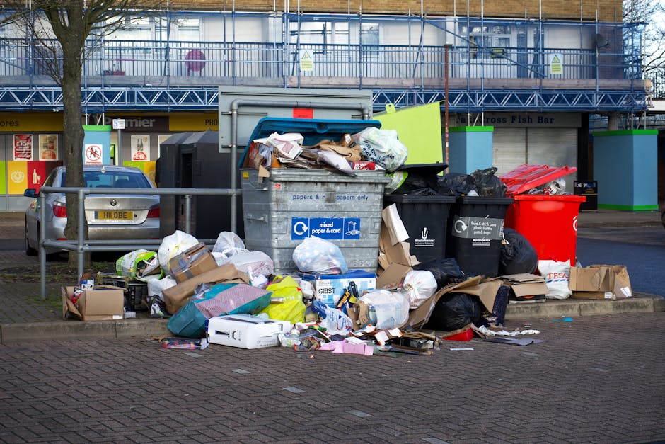 An alleyway scene showing a large, black plastic roll of rubbish, positioned in the foreground on the uneven, stained pavement. The alley is flanked by narrow walls of adjacent buildings, with the lef